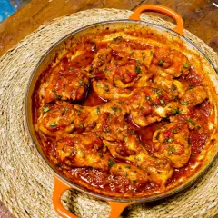 Sabroso pollo al chilindrón en cazuela naranja, servido sobre una esterilla de mimbre en una mesa de madera rústica.