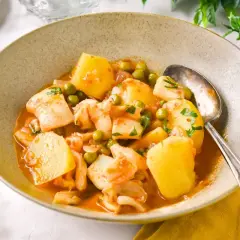 Un tazón de sepia con patatas, guisantes y perejil picado, servido con una cuchara plateada.