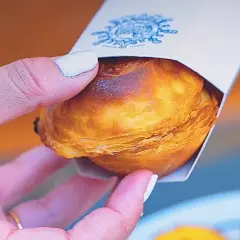 Mano sosteniendo un pastelito de Belém recién horneado, con su masa hojaldrada dorada y un toque de caramelo.