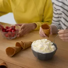 Dos personas preparan helado casero. Hay un tazón de helado cremoso, fresas, conos de helado y un vaso.