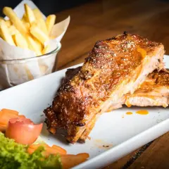Costillas al horno jugosas y doradas, servidas con papas fritas crujientes y una guarnición fresca de lechuga, zanahoria y tomate.