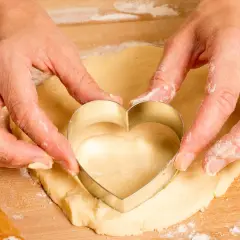 Manos cortan masa de galletas con un cortador en forma de corazón.