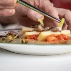 Manos preparan pa amb oli con queso, tomate, aceitunas y alcachofas en una tostada.