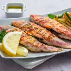 Cuatro salmonetes al horno, dorados y jugosos, servidos con rodajas de limón, perejil y patatas fritas.