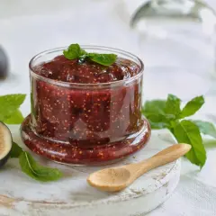 Tarro de mermelada de higos casera, con higos frescos y hojas de menta alrededor. Una cuchara de madera descansa cerca.