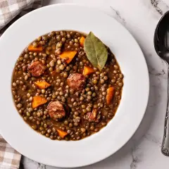 Plato de lentejas de bote con chorizo y zanahoria, decorado con una hoja de laurel. Una cuchara plateada descansa a la derecha.