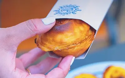 Mano sosteniendo un pastelito de Belém recién horneado, con su masa hojaldrada dorada y un toque de caramelo.