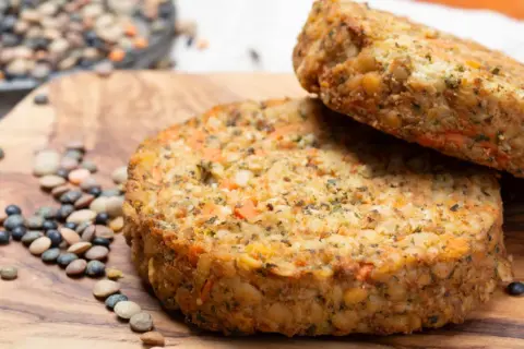 Dos hamburguesas de lentejas caseras, con trozos de zanahoria y hierbas, reposan sobre una tabla de madera junto a lentejas crudas.