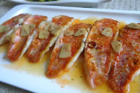 Deliciosos salmonetes a la plancha con láminas de ajo y un toque de guindilla, servidos en una salsa dorada.