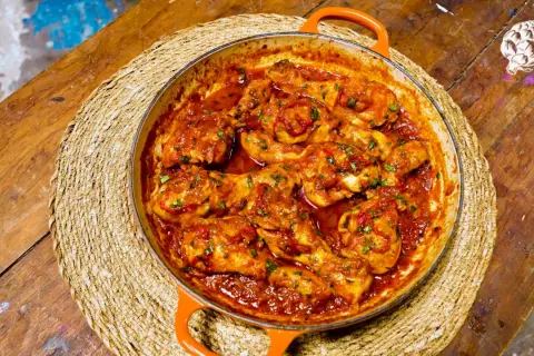 Sabroso pollo al chilindrón en cazuela naranja, servido sobre una esterilla de mimbre en una mesa de madera rústica.