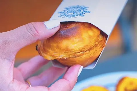 Mano sosteniendo un pastelito de Belém recién horneado, con su masa hojaldrada dorada y un toque de caramelo.