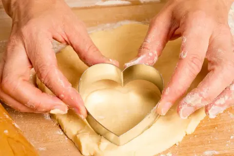 Manos cortan masa de galletas con un cortador en forma de corazón.