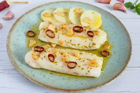 Dos lomos de merluza a la bilbaina, con patatas, ajo laminado y guindilla, servidos en plato azul.