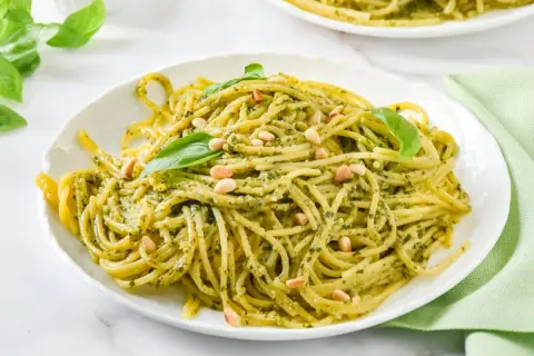 Espaguetis al pesto, cubiertos con piñones y hojas de albahaca fresca, servidos en un plato blanco.