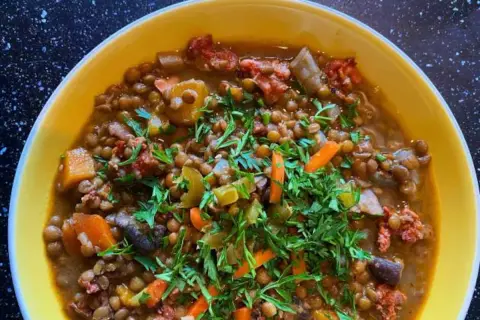Un reconfortante potaje de lentejas con trozos de chorizo, zanahoria y perejil fresco, servido en un cuenco amarillo.