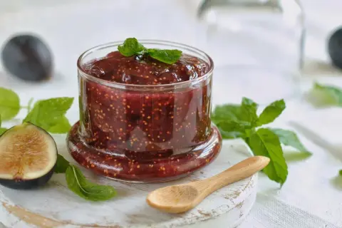 Tarro de mermelada de higos casera, con higos frescos y hojas de menta alrededor. Una cuchara de madera descansa cerca.