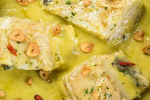 Merluza al pil pil, con trozos de pescado blanco en una salsa amarilla cremosa, adornada con láminas de ajo crujiente y guindillas.
