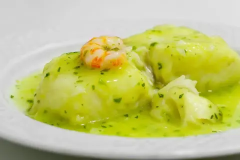Merluza en salsa verde, un plato tradicional con trozos de pescado blanco y un camarón encima, bañado en una deliciosa salsa verde.