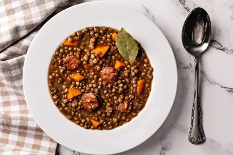Plato de lentejas de bote con chorizo y zanahoria, decorado con una hoja de laurel. Una cuchara plateada descansa a la derecha.