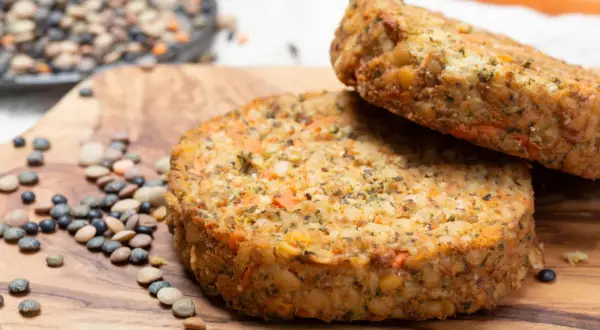 Dos hamburguesas de lentejas caseras, con trozos de zanahoria y hierbas, reposan sobre una tabla de madera junto a lentejas crudas.