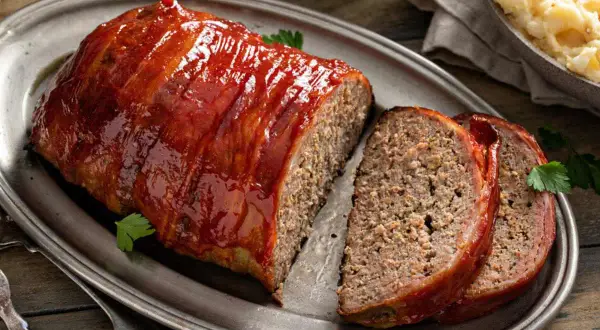Un jugoso pastel de carne cubierto de glaseado rojo brillante, envuelto en tocino y servido en una bandeja de plata.