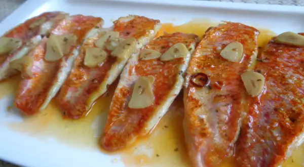 Deliciosos salmonetes a la plancha con láminas de ajo y un toque de guindilla, servidos en una salsa dorada.