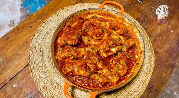 Sabroso pollo al chilindrón en cazuela naranja, servido sobre una esterilla de mimbre en una mesa de madera rústica.