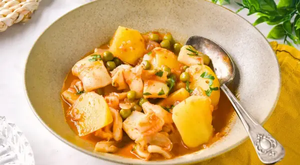 Un tazón de sepia con patatas, guisantes y perejil picado, servido con una cuchara plateada.