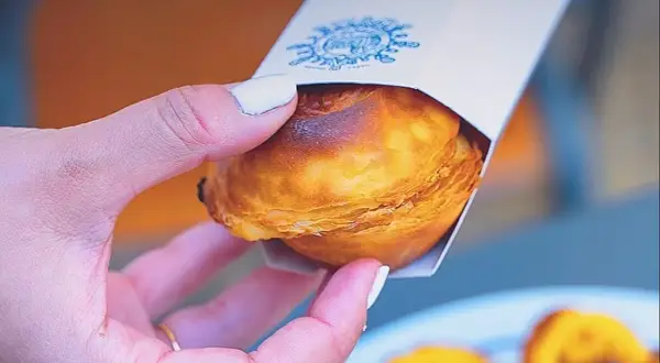 Mano sosteniendo un pastelito de Belém recién horneado, con su masa hojaldrada dorada y un toque de caramelo.