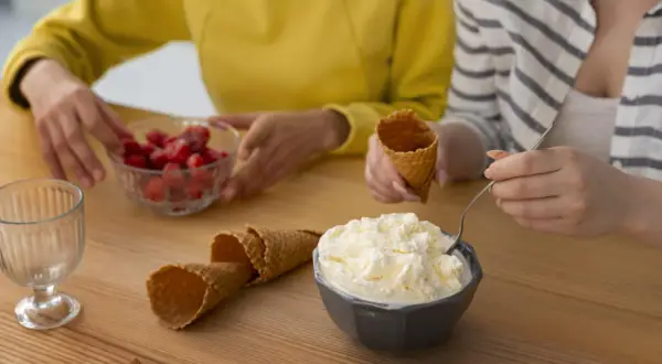 Dos personas preparan helado casero. Hay un tazón de helado cremoso, fresas, conos de helado y un vaso.
