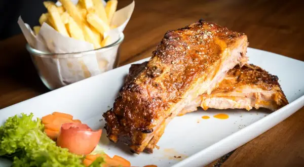 Costillas al horno jugosas y doradas, servidas con papas fritas crujientes y una guarnición fresca de lechuga, zanahoria y tomate.