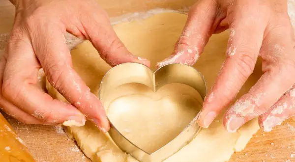 Manos cortan masa de galletas con un cortador en forma de corazón.