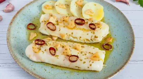 Dos lomos de merluza a la bilbaina, con patatas, ajo laminado y guindilla, servidos en plato azul.