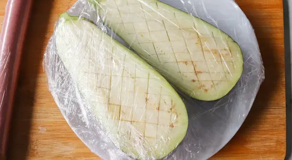Dos mitades de berenjena cortadas en cubos y cubiertas con plástico, listas para cocinar berenjenas al microondas.