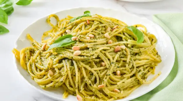 Espaguetis al pesto, cubiertos con piñones y hojas de albahaca fresca, servidos en un plato blanco.