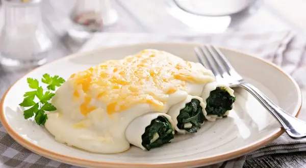 Canelones de espinacas cubiertos con bechamel y queso gratinado, servidos en un plato con perejil fresco.