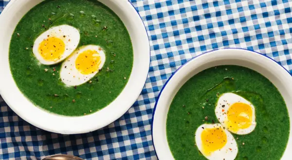 Dos cuencos de espinacas a la crema con huevos duros cortados por la mitad, servidos sobre un mantel de cuadros azules y blancos.