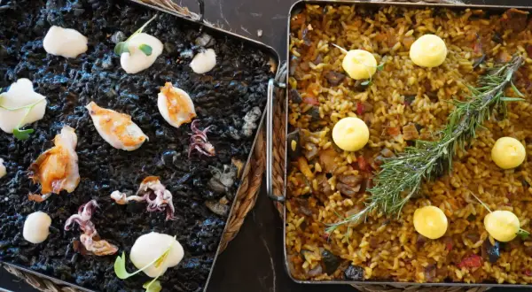 Dos arroces a la llauna: uno negro con calamares y alioli, y otro dorado con verduras y romero.