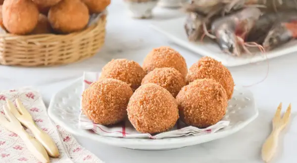 Croquetas de gambas doradas y crujientes en un plato, con gambas crudas al fondo.