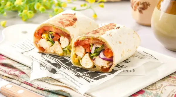 Dos burritos de pollo cortados por la mitad, rellenos de pollo, tomate, lechuga y cebolla morada, servidos sobre papel de periódico.