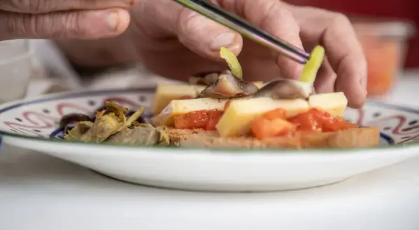 Manos preparan pa amb oli con queso, tomate, aceitunas y alcachofas en una tostada.