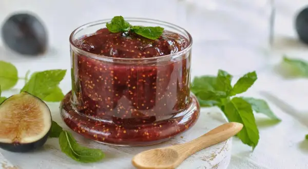 Tarro de mermelada de higos casera, con higos frescos y hojas de menta alrededor. Una cuchara de madera descansa cerca.