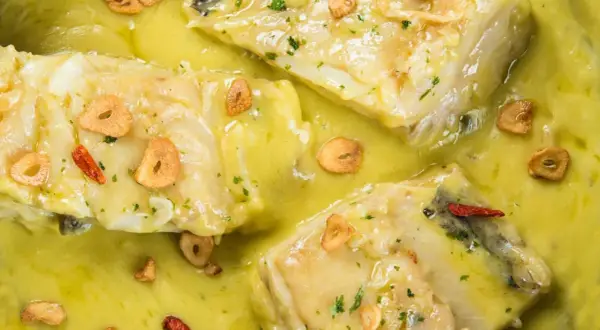 Merluza al pil pil, con trozos de pescado blanco en una salsa amarilla cremosa, adornada con láminas de ajo crujiente y guindillas.