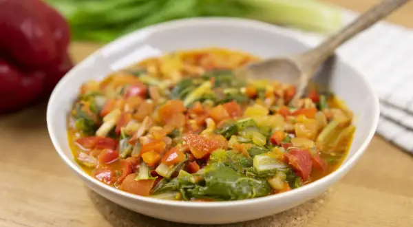 Un plato humeante de guiso de verduras, ideal para recetas con acelgas. Contiene trozos de tomate, pimiento rojo y hojas verdes.