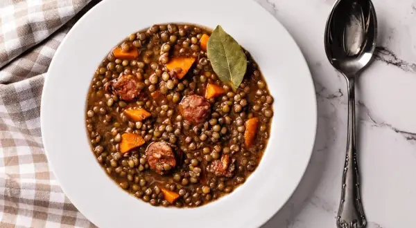 Plato de lentejas de bote con chorizo y zanahoria, decorado con una hoja de laurel. Una cuchara plateada descansa a la derecha.