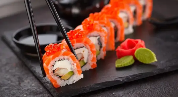 Palillos recogen un rollo de sushi con salmón y huevas de salmón, listo para disfrutar. Aprende como hacer sushi.