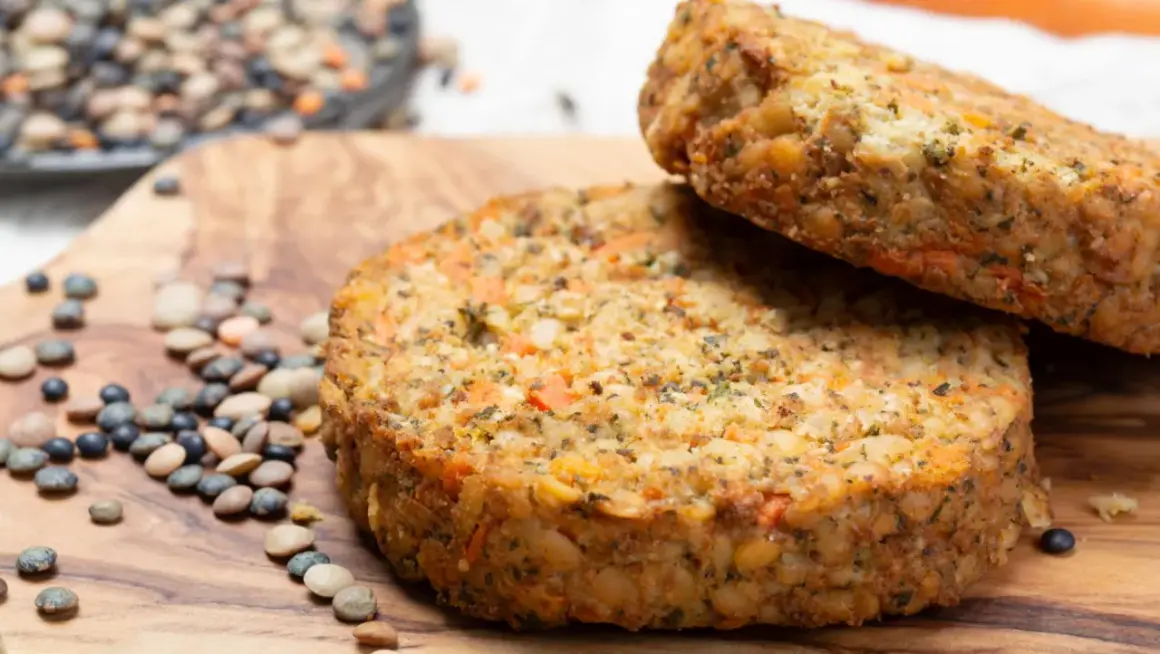 Dos hamburguesas de lentejas caseras, con trozos de zanahoria y hierbas, reposan sobre una tabla de madera junto a lentejas crudas.