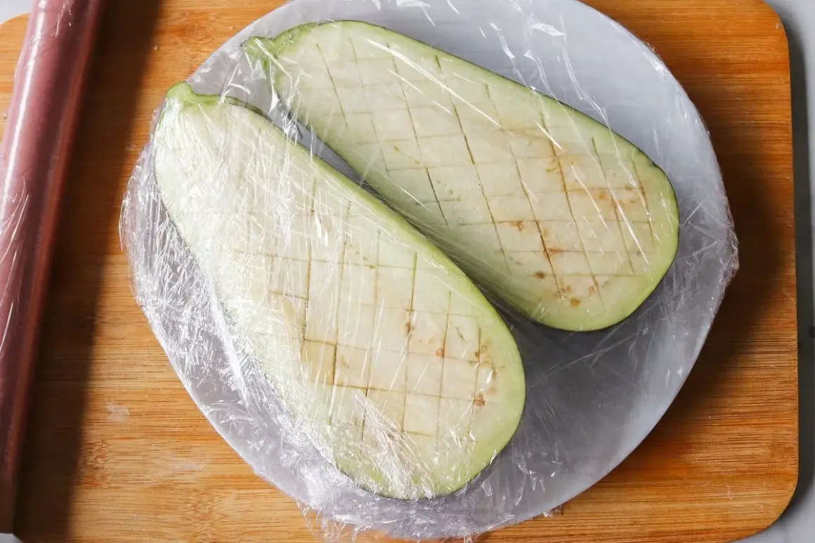 Dos mitades de berenjena cortadas en cubos y cubiertas con plástico, listas para cocinar berenjenas al microondas.