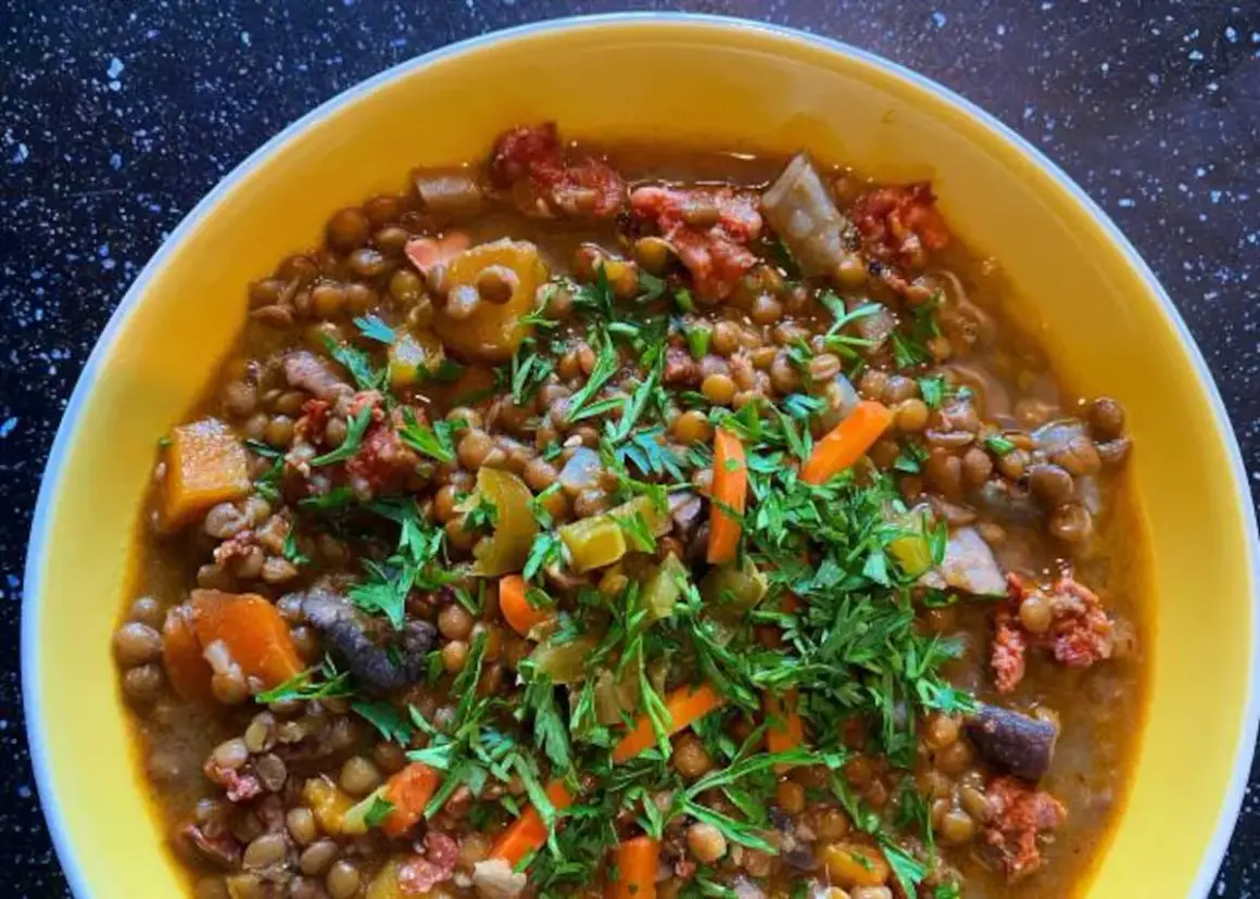 Un reconfortante potaje de lentejas con trozos de chorizo, zanahoria y perejil fresco, servido en un cuenco amarillo.