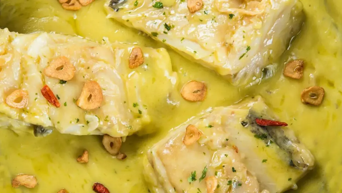 Merluza al pil pil, con trozos de pescado blanco en una salsa amarilla cremosa, adornada con láminas de ajo crujiente y guindillas.